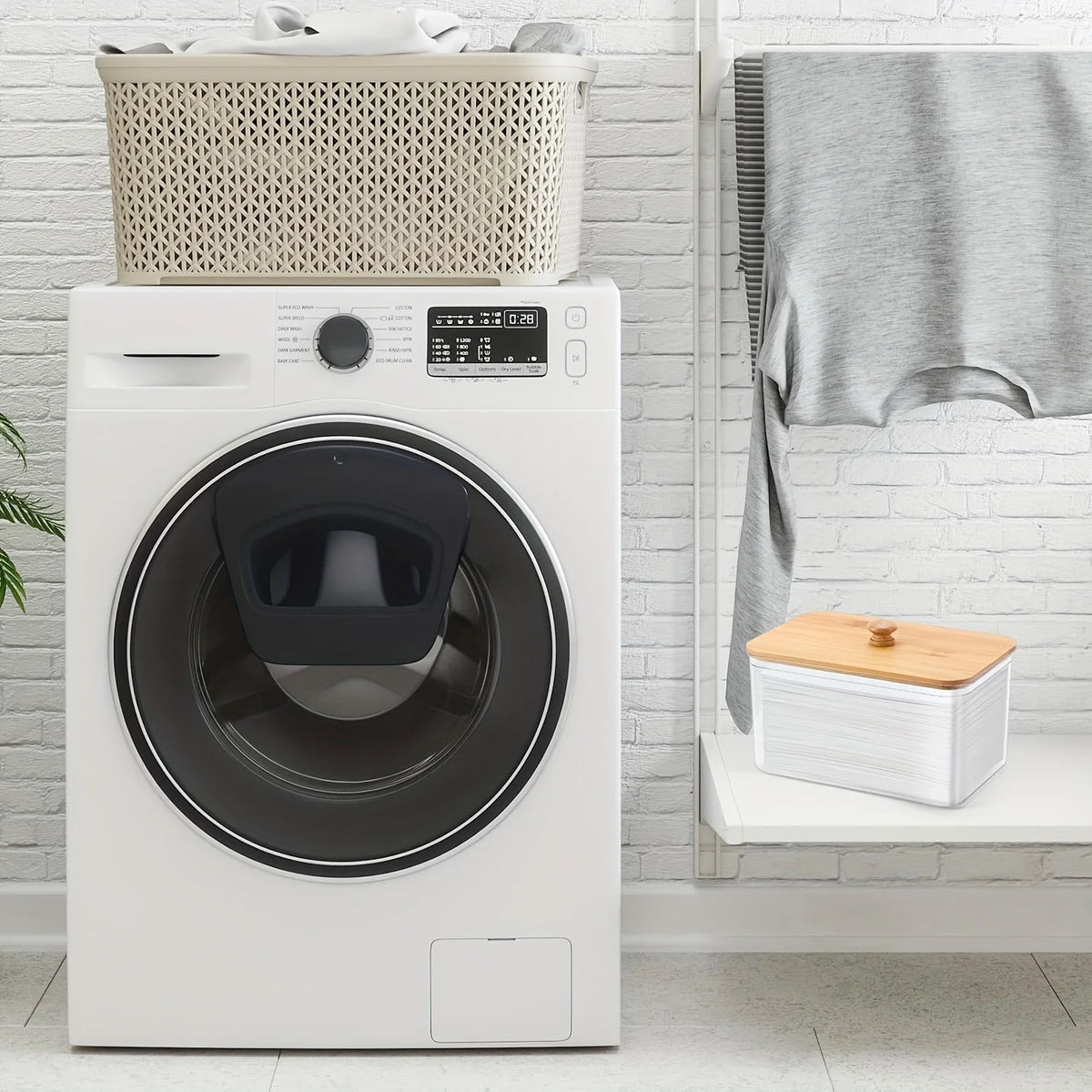 A white washing machine is shown next to a woven basket and a gray towel hanging on a white brick wall.