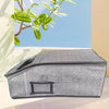 A gray foldable fabric hat storage box with a zipper is shown on a light surface next to a green branch.