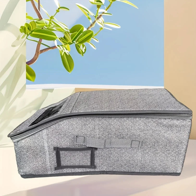 A gray foldable fabric hat storage box with a zipper is shown on a light surface next to a green branch.
