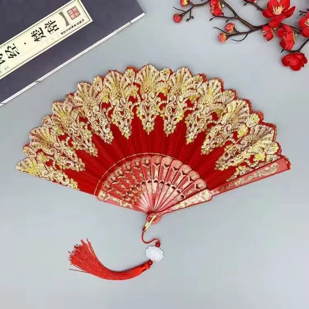 A red and gold folding hand fan with intricate patterns, displayed on a gray surface.