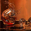 A glass of amber liquid is being poured into a bowl, creating splashing water and ice cubes on a reflective surface.