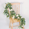 A wooden chair decorated with a garland of artificial eucalyptus leaves and white roses.