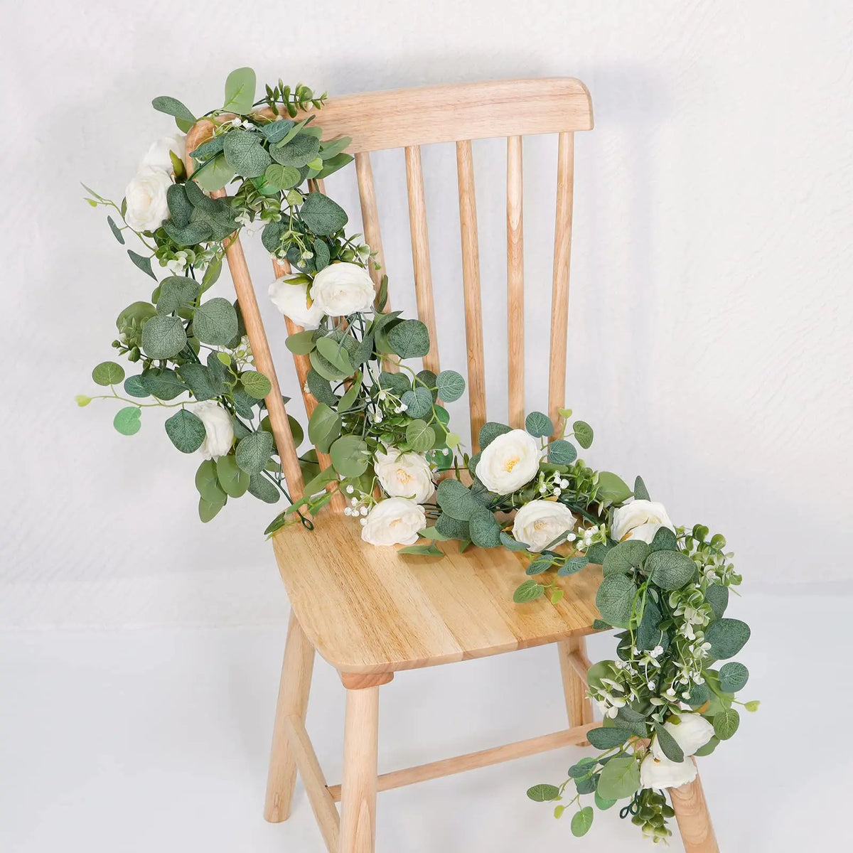 A wooden chair decorated with a garland of artificial eucalyptus leaves and white roses.