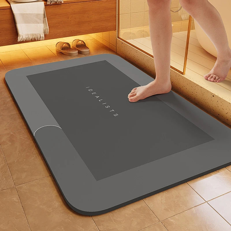A gray non-slip bath mat with the word IDEALISTS embossed on it is placed on a tiled floor.