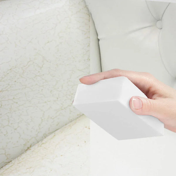 A hand holds a white melamine cleaning sponge, often called a magic eraser, against a white upholstered surface.