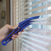 A hand is using a blue reusable household cleaning brush to clean slats on a window blind.