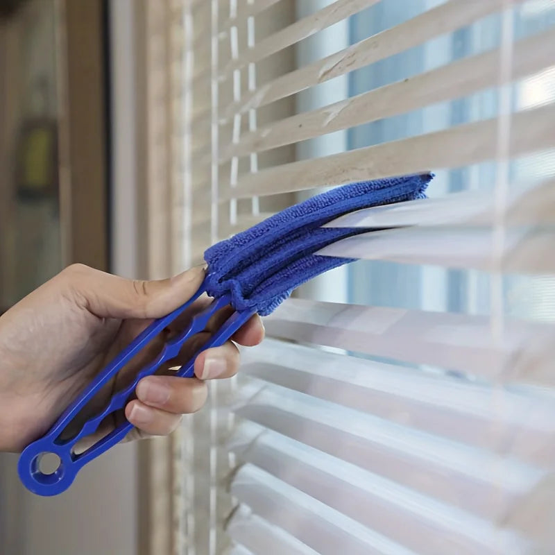 A hand is using a blue reusable household cleaning brush to clean slats on a window blind.