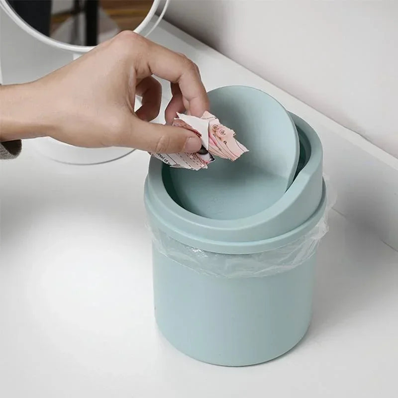 A hand is placing crumpled tissue paper into the flip cover of a light blue compact desktop trash can.