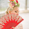 A woman holds a red folding fan with intricate gold embroidery against a bright background.