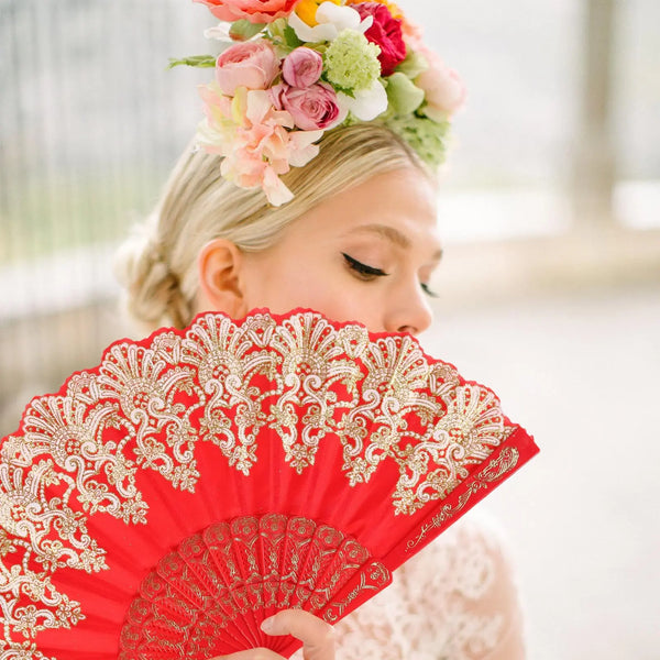 A woman holds a red folding fan with intricate gold embroidery against a bright background.