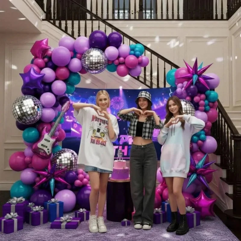 Drei junge Frauen posieren vor einem lila und türkis verzierten Ballonbogen mit silbernen und lila Ballons.