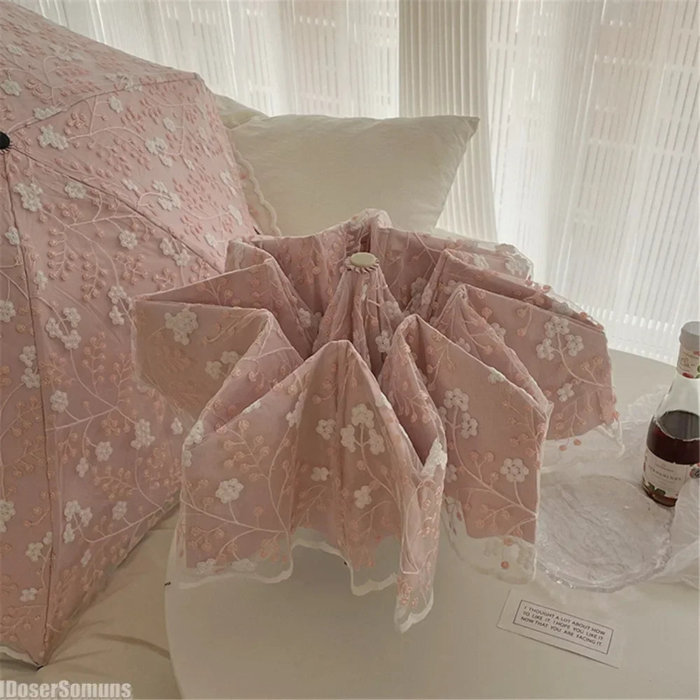 A pink full-automatic embroidered sun umbrella is displayed on a white surface next to sheer curtains.