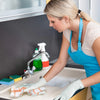 A woman wearing a blue apron and white gloves is cleaning a kitchen sink with a faucet and green dish soap.