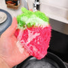 A hand holds a bright green and pink strawberry scrubber sponge for kitchen cleaning.
