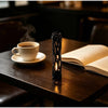 A dark wooden table features a cup of coffee and an illuminated willow stick night light projector next to an open book.