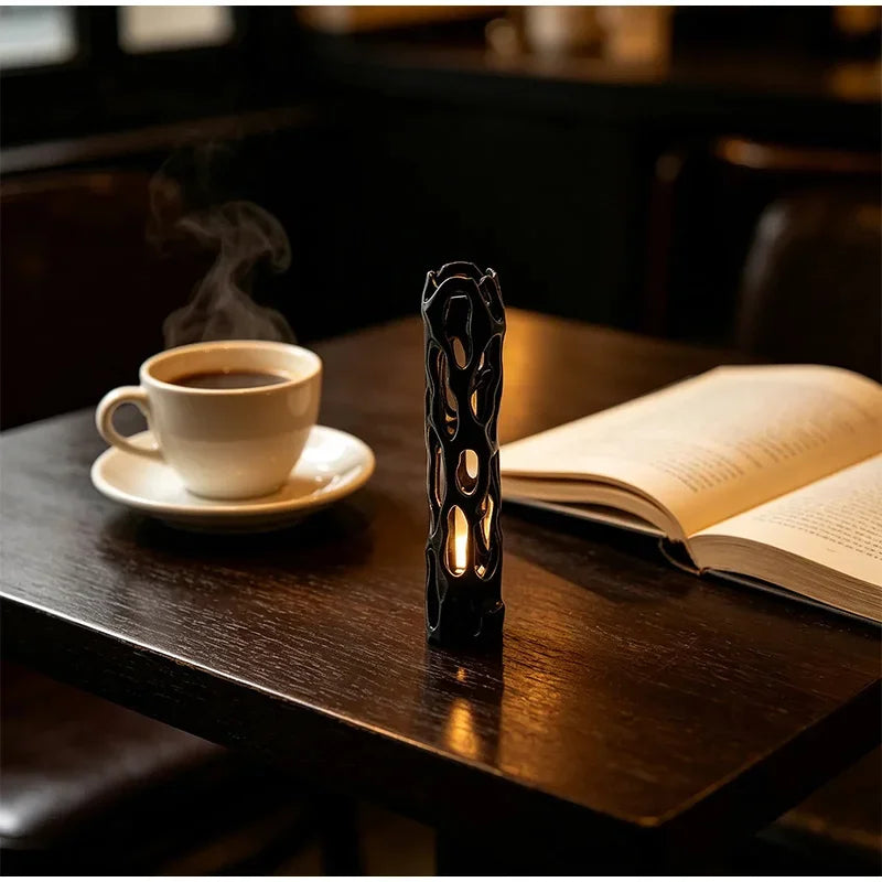 A dark wooden table features a cup of coffee and an illuminated willow stick night light projector next to an open book.