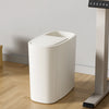 A white odor-sealing trash can with a press lid sits on a wooden floor next to a dispenser.