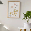A framed print featuring delicate, watercolor-style butterflies in shades of orange, gold, and gray on a white wall.