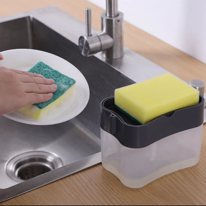 A hand is pressing a yellow sponge onto a plate in a stainless steel sink next to a clear dispenser.