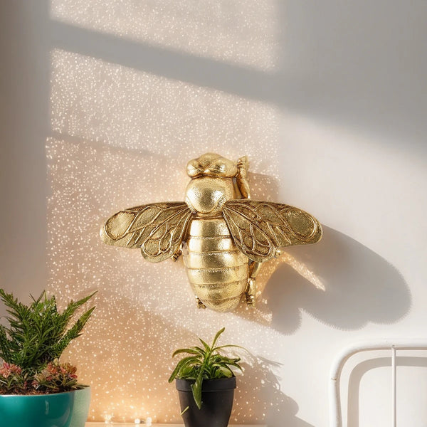 A golden bee wall decor hanging on a white wall next to potted plants.