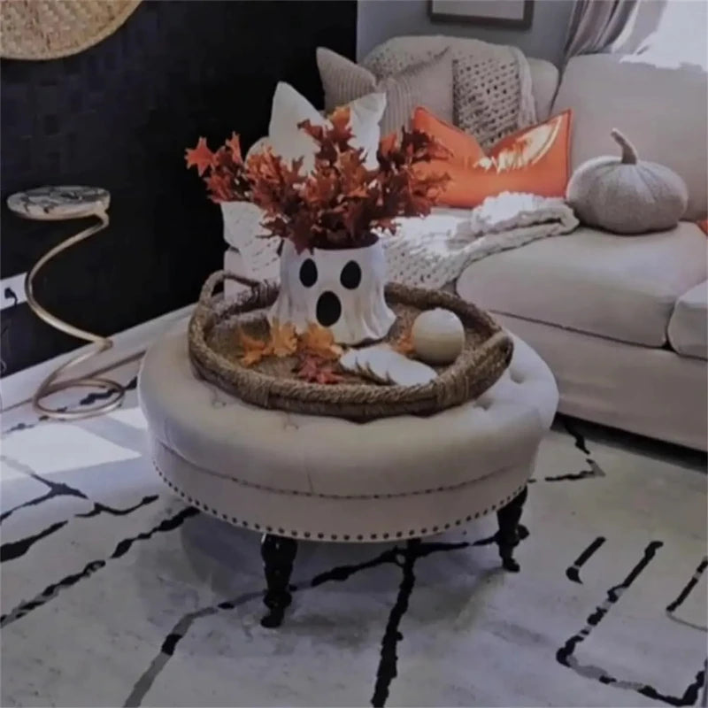 Un vase fantôme en céramique fait main avec un centre de citrouille, posé sur un tabouret rembourré dans un salon chaleureux.