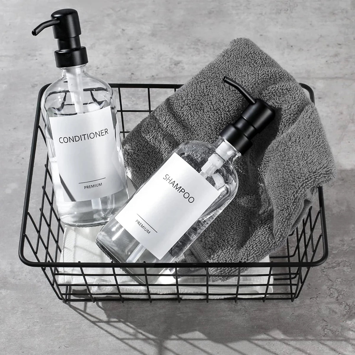 A black wire basket holds two clear glass bottles of premium conditioner and shampoo with stainless steel pumps resting on a 