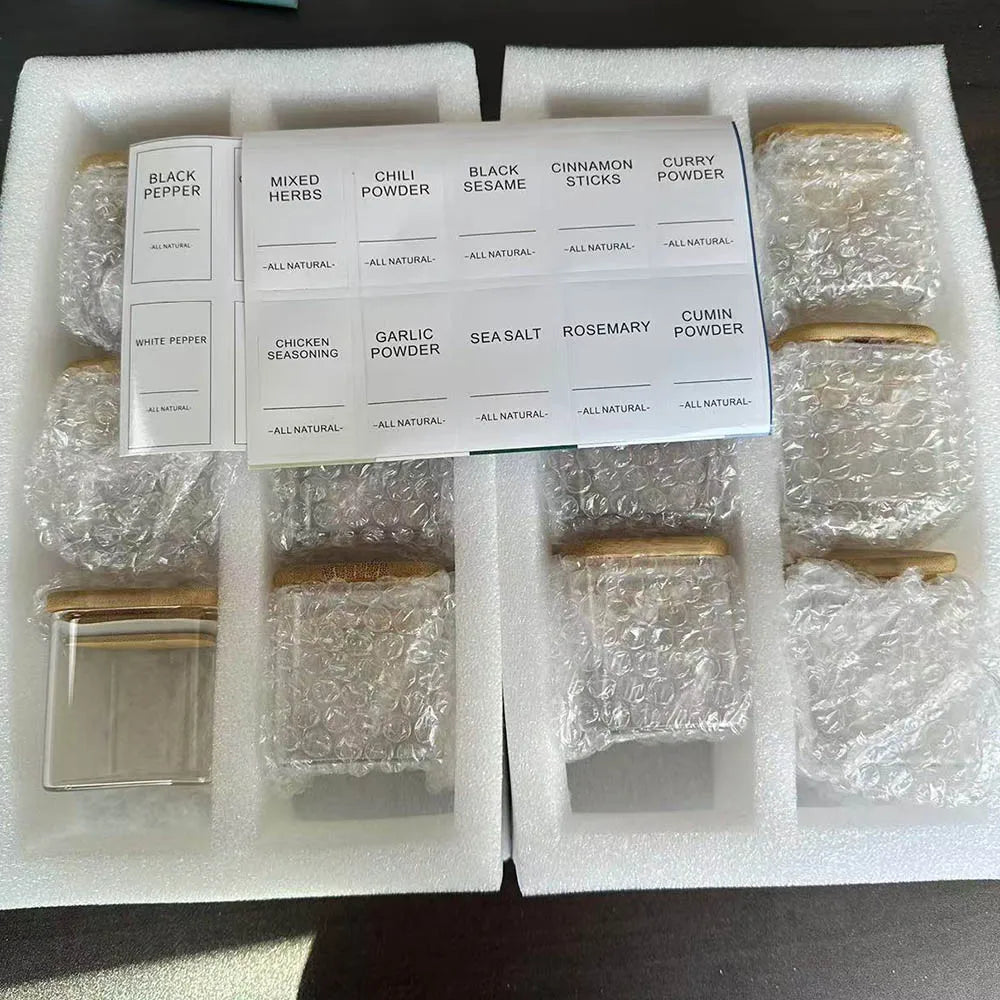 A white box contains five glass spice jars with bamboo lids labeled for various spices like black pepper chili powder and cin