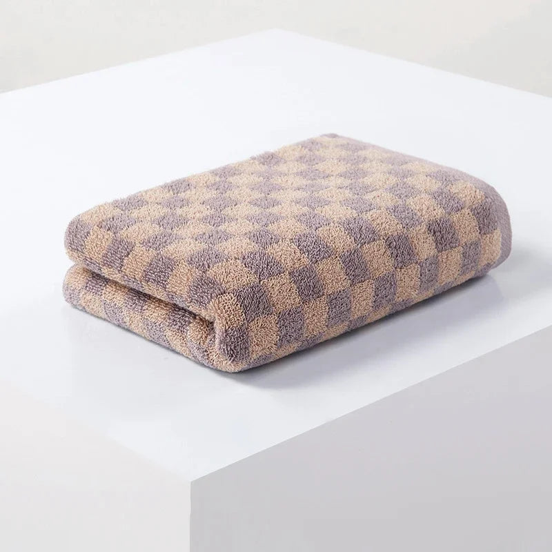 A folded plaid cotton bath towel set with a beige and purple checkered pattern resting on a white surface.