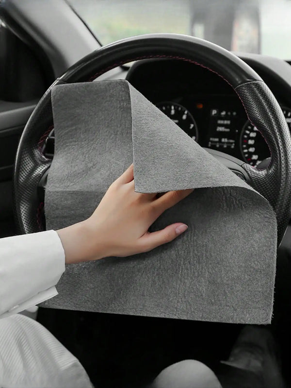 A hand is holding a gray reusable microfiber cleaning cloth on the steering wheel of a car.