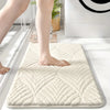 A beige plush non-slip bathroom rug is placed on a white floor next to a person's leg.