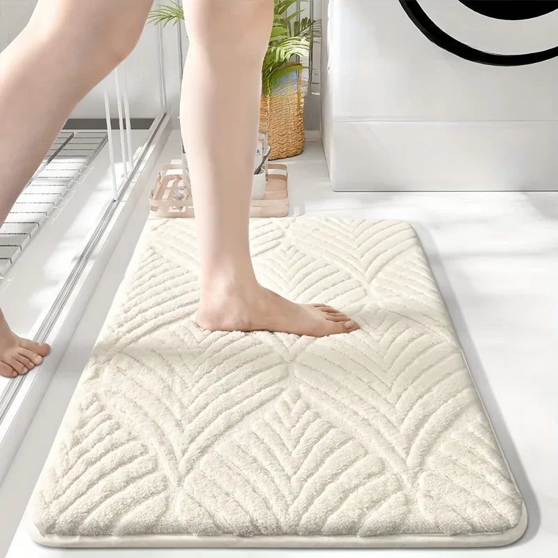 A beige plush non-slip bathroom rug is placed on a white floor next to a person's leg.