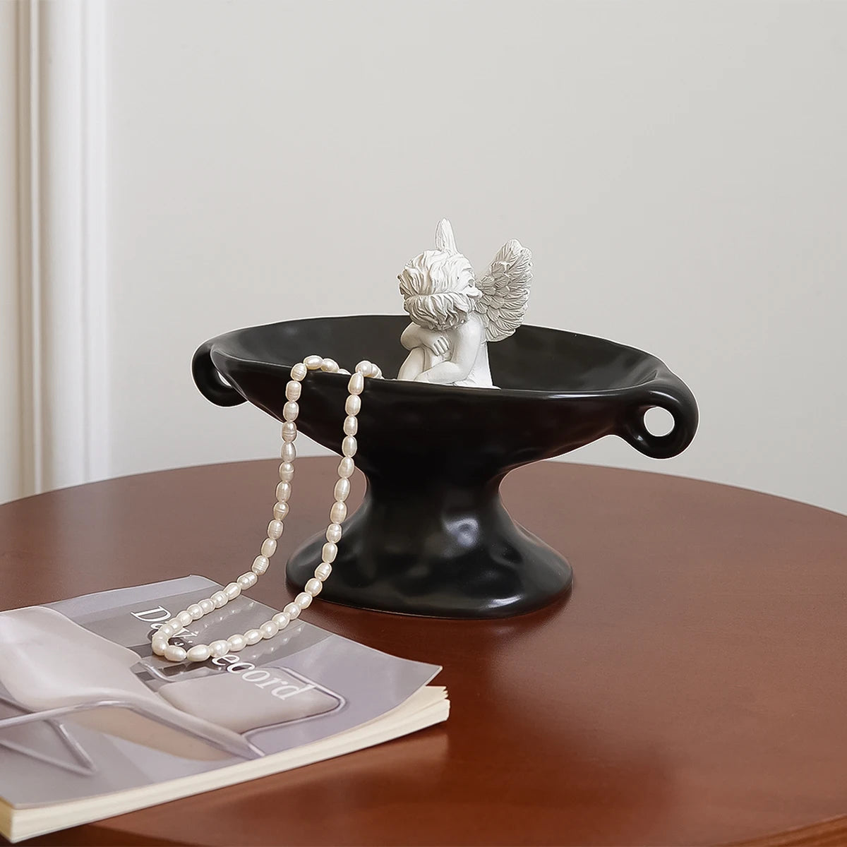 A black ceramic double-ear soap dish with a white angel figurine sits on a wooden table.