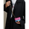 A hand holds a purple magnetic crossbody bag with a letter flap attached to a gold chain against a black coat.