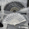 A delicate white lace folding hand fan with a gray edge and gold accents displayed on sheer fabric.
