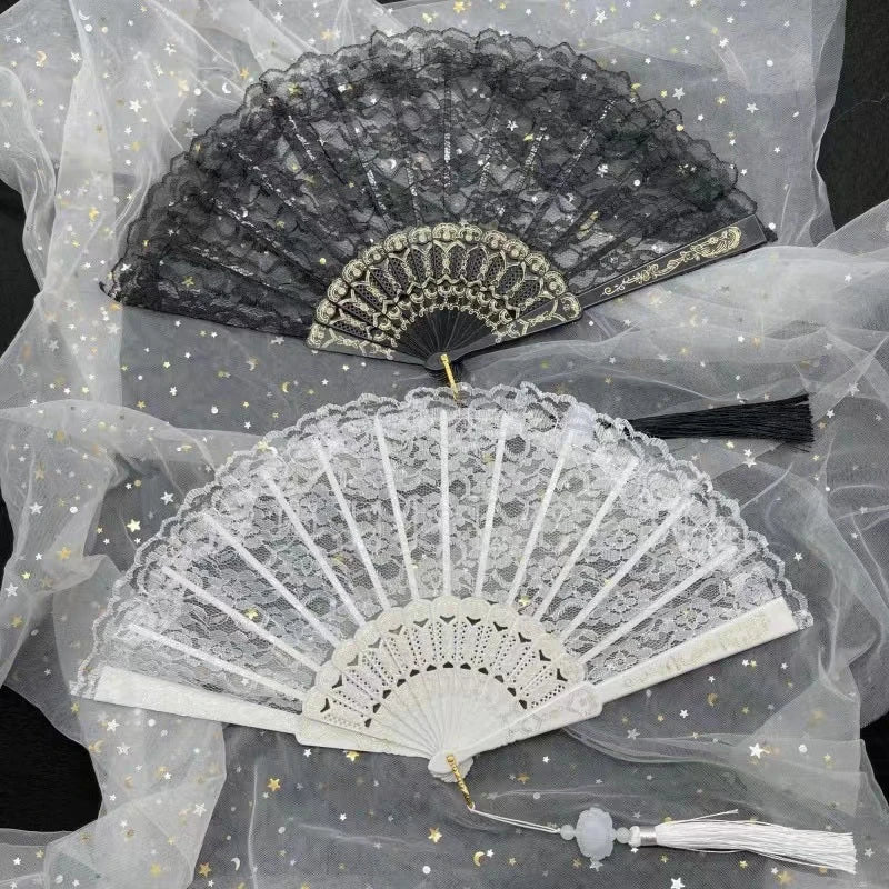 A delicate white lace folding hand fan with a gray edge and gold accents displayed on sheer fabric.