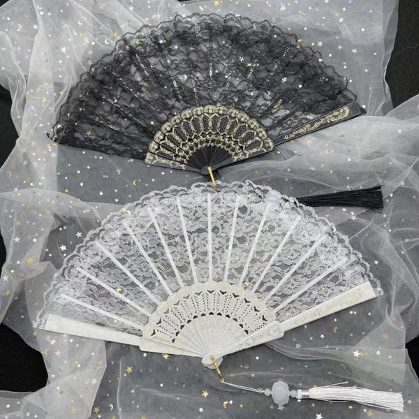 A delicate white lace folding hand fan with a gray edge and gold accents displayed on sheer fabric.