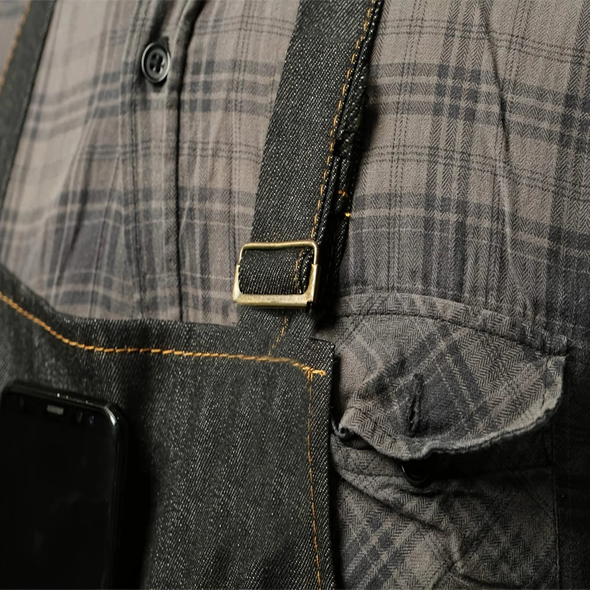 Close-up of a dark gray plaid denim tool apron with a leather buckle and pocket on the chest.