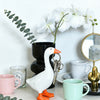 A white goose sculpture with an orange beak and legs is displayed next to a key chain and white flowers on a light surface.
