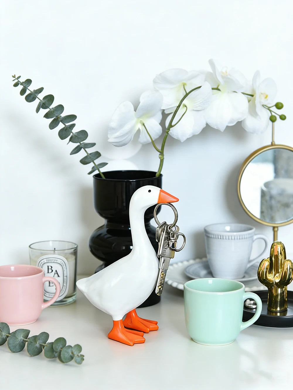 A white goose sculpture with an orange beak and legs is displayed next to a key chain and white flowers on a light surface.