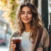 A young woman holding a dark coffee cup outdoors, wearing a knitted cardigan.