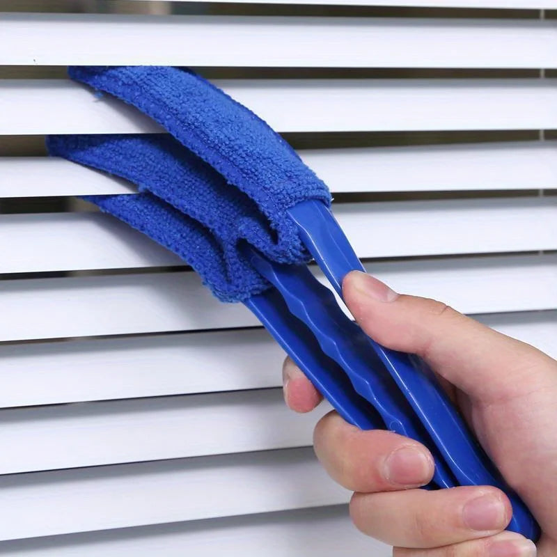 A hand holds a blue reusable household cleaning brush with a blue cleaning head against a white slatted surface.
