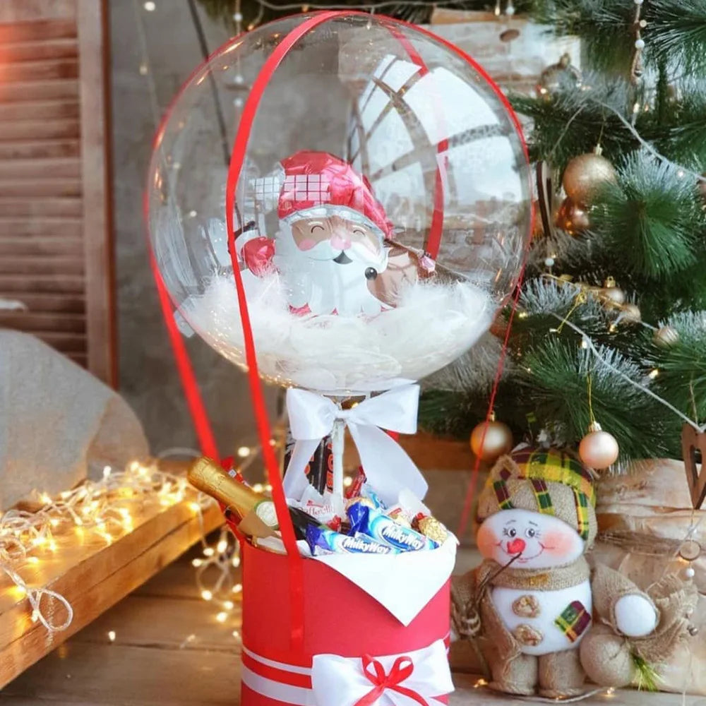 A clear latex balloon filled with a Santa face decoration tied to a red ribbon and placed next to a Christmas tree.