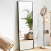 A full body standing wall mirror with a black frame reflecting a potted palm plant and a beige sofa.