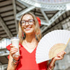 Une femme souriante tient une boisson blanche et un grand éventail décoratif blanc contre un arrière-plan architectural flou.