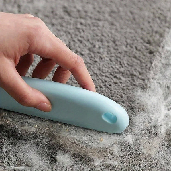 A hand is using a light blue pet hair remover brush on a gray, fuzzy surface.