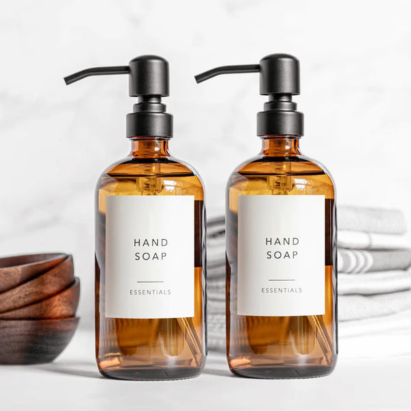 Two amber glass hand soap dispensers with black pumps and essential oils are displayed on a white surface.