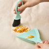 A hand is holding a light blue mini broom and dustpan set on a beige surface.