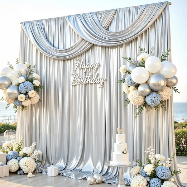 A silver satin shimmer backdrop with draped curtains and floral arrangements for a birthday event.