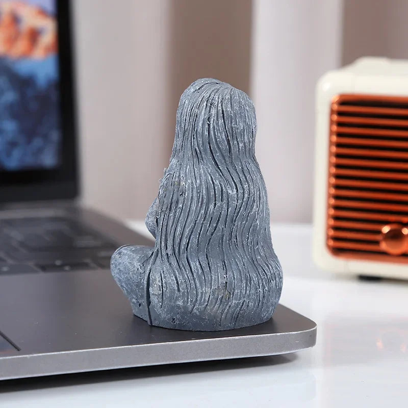 A gray resin statue of a long, wavy-haired figure in a meditative pose on a dark surface.