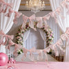 A pink decorative bowknot banner garland set hangs in a wedding or event setting surrounded by white drapery and floral arran